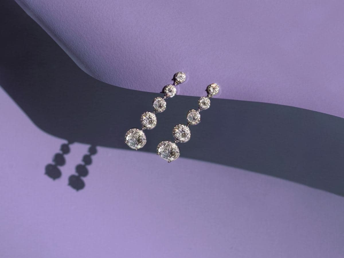 Pair of graduated five-stone drops, each round stone in a thin halo, white metal, hanging over a lavender paper edge