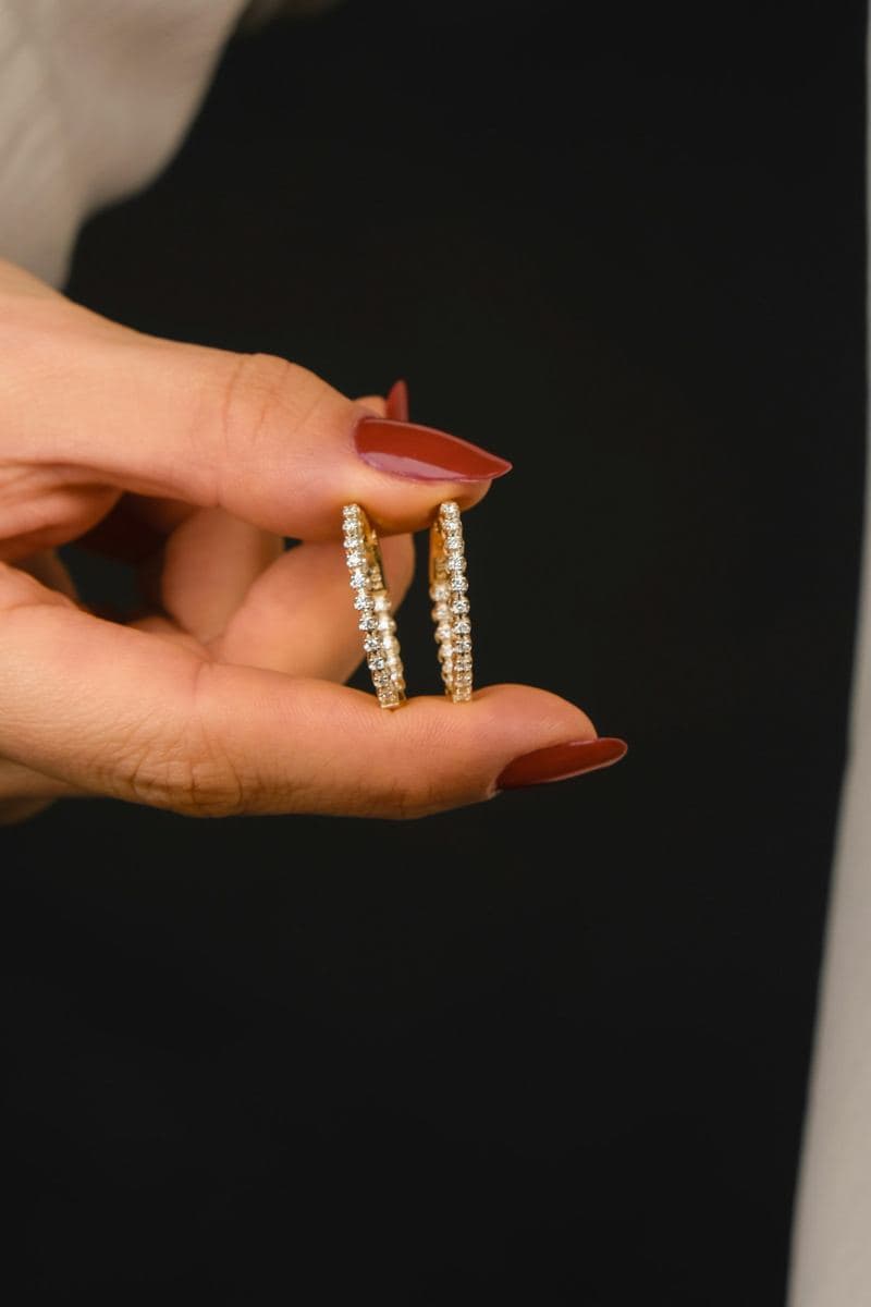 Hand with red nails holding a pair of slim inside-out hoops set with round stones in yellow gold