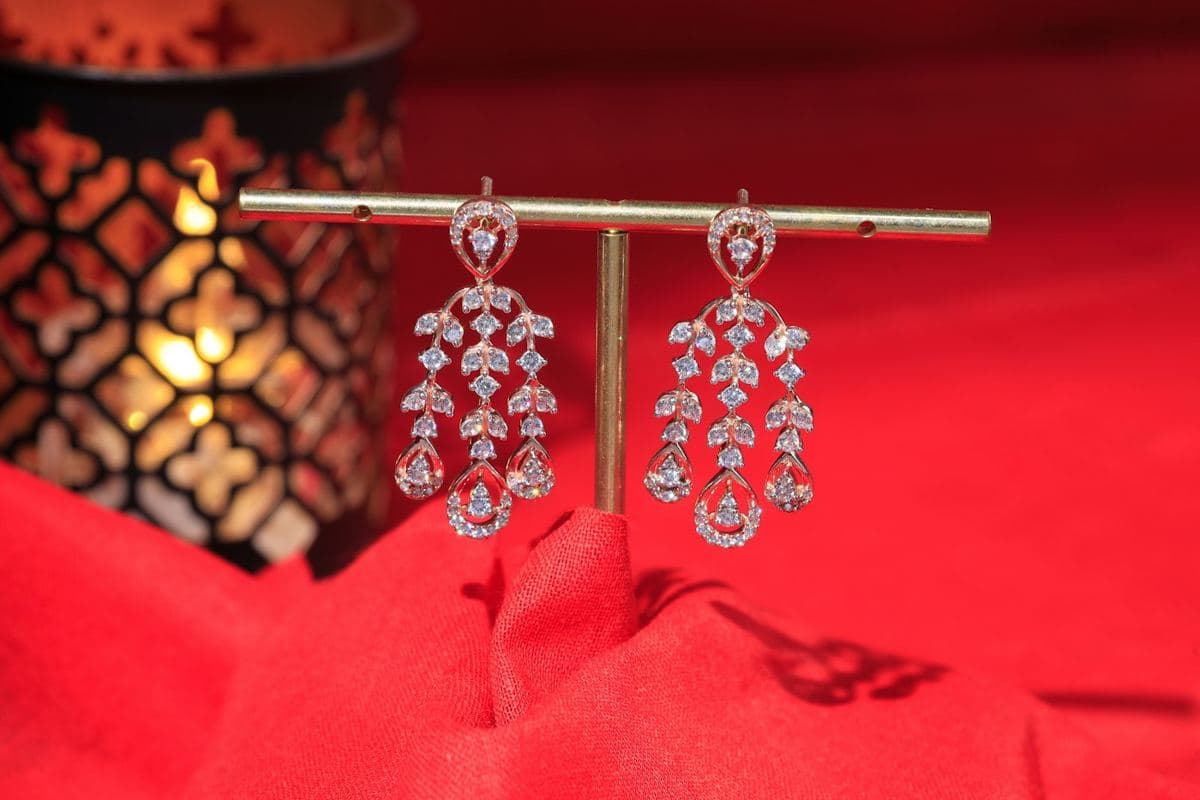 Pair of chandelier earrings of leaf-patterned round stones ending in teardrop frames, rose-gold tone, hung on a brass bar stand