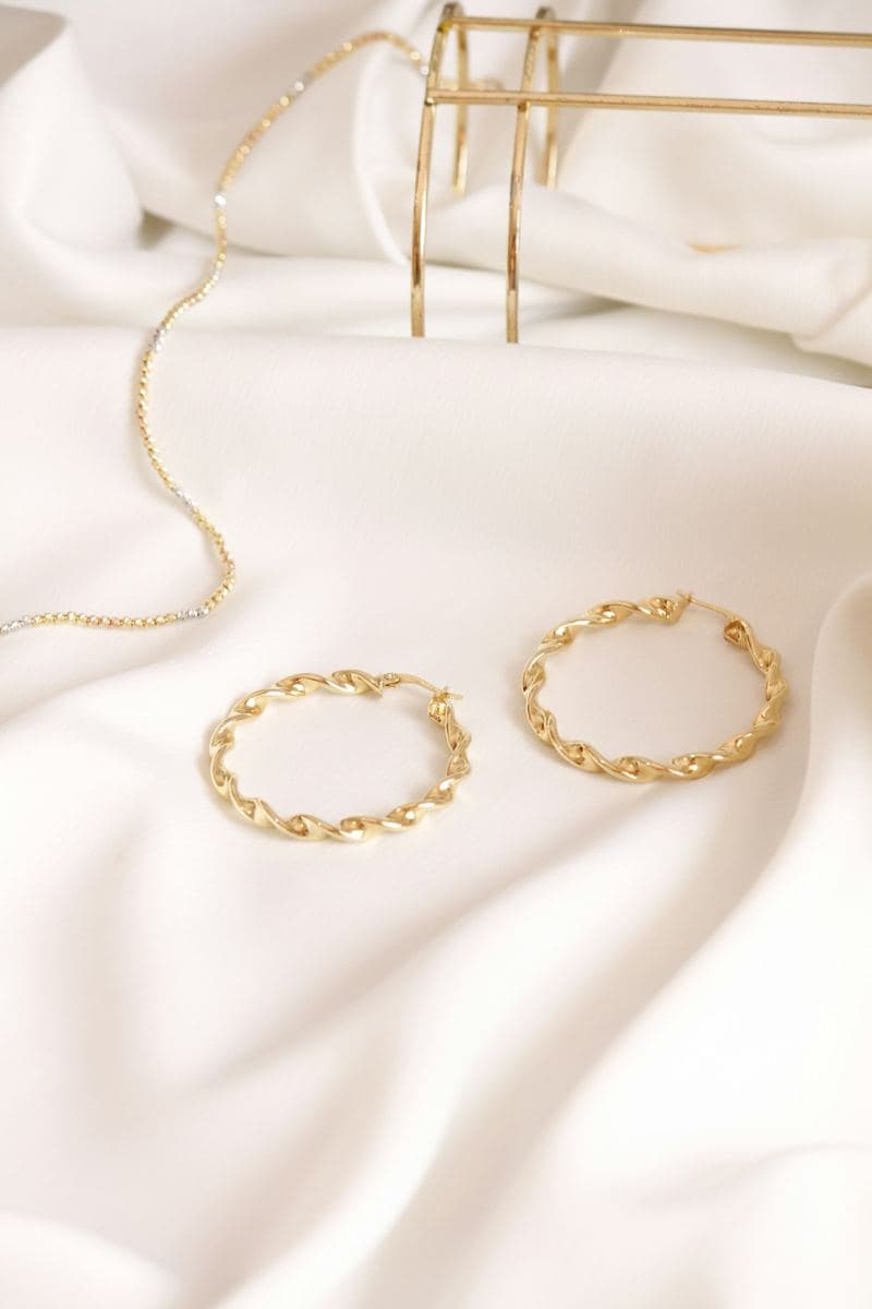 Pair of twisted yellow-gold hoops with hinged clasps on white satin, a chain blurred behind