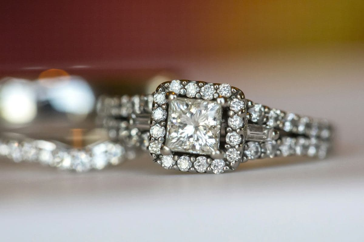 Princess center in a square stone halo with baguette side stones on a split pavé shank; a curved stone band blurred behind