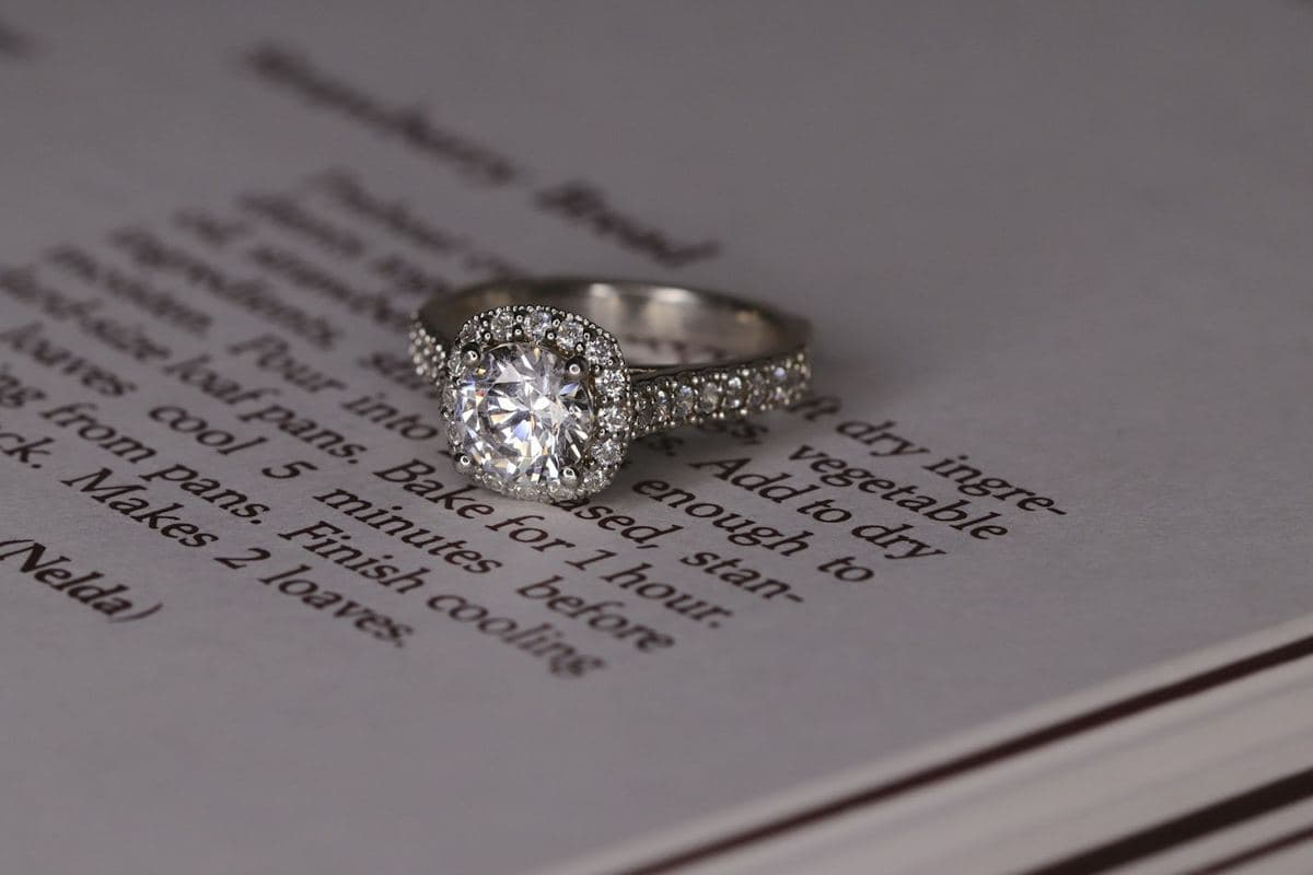 Round center in a cushion-shaped milgrain halo on a pavé white-metal band, resting on a printed recipe page