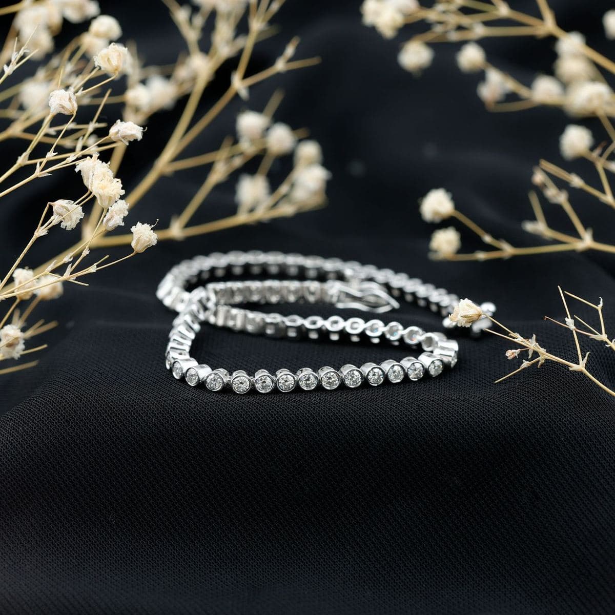 A white-metal bezel-set tennis bracelet of uniform round stones, coiled on black fabric with its box clasp visible, framed by dried white flowers