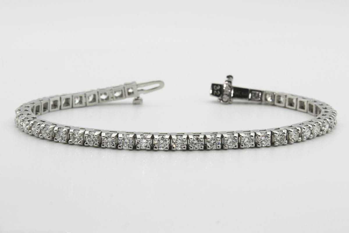 A single-row white-metal tennis bracelet of uniform round stones in four-prong settings, laid on light grey with its box clasp open at the top