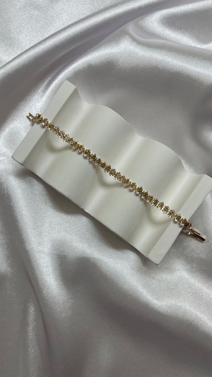 A yellow-gold tennis bracelet of uniform marquise-shaped white stones set side-on, with a fold-over clasp, laid across a white wavy tray on satin