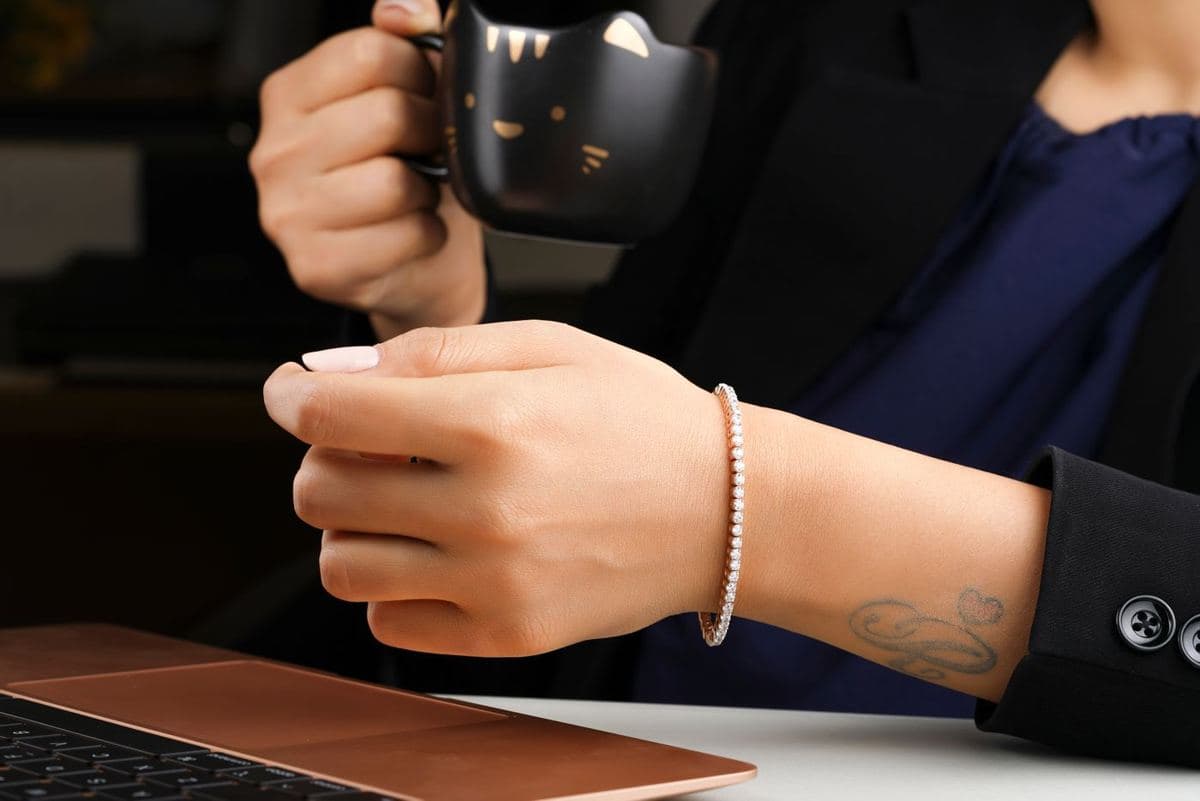 A woman's wrist (no face) wearing a thin rose-gold line of uniform small round white stones, holding a black mug beside a laptop