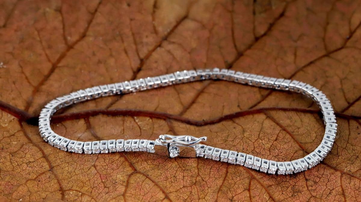 A closed loop of uniform prong-set round white stones in white metal with a box clasp and safety latch, lying on a dried leaf