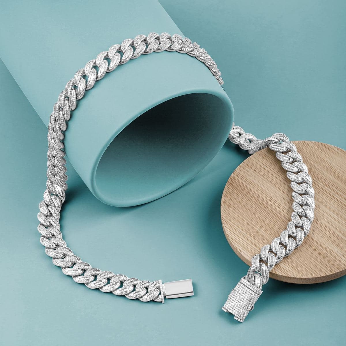Flat-lay of a white-metal cuban link chain with baguette-set link centers edged in small round stones, pavé box clasp, draped over a teal tube and wooden disc