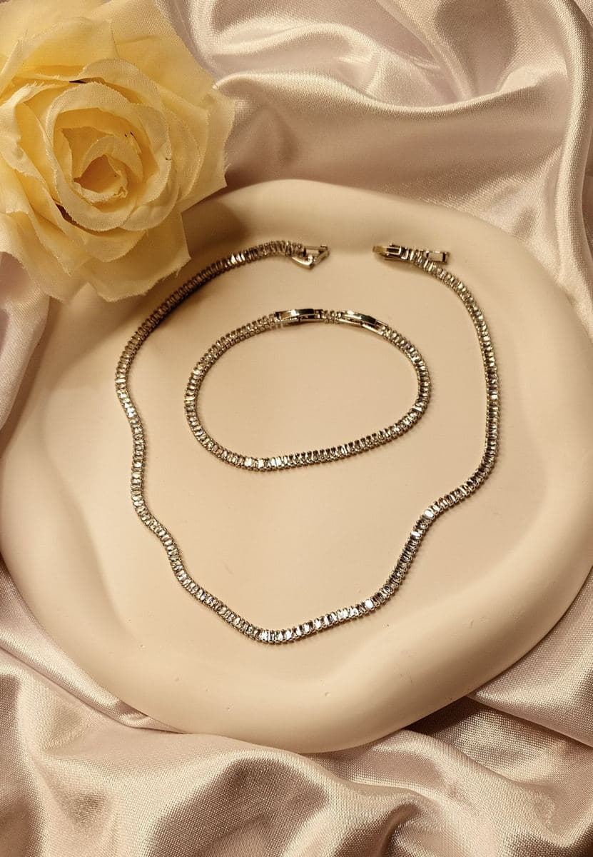 Flat lay of a white-metal baguette-stone tennis necklace with box clasp, with a matching baguette tennis bracelet coiled inside it, on a cream tray