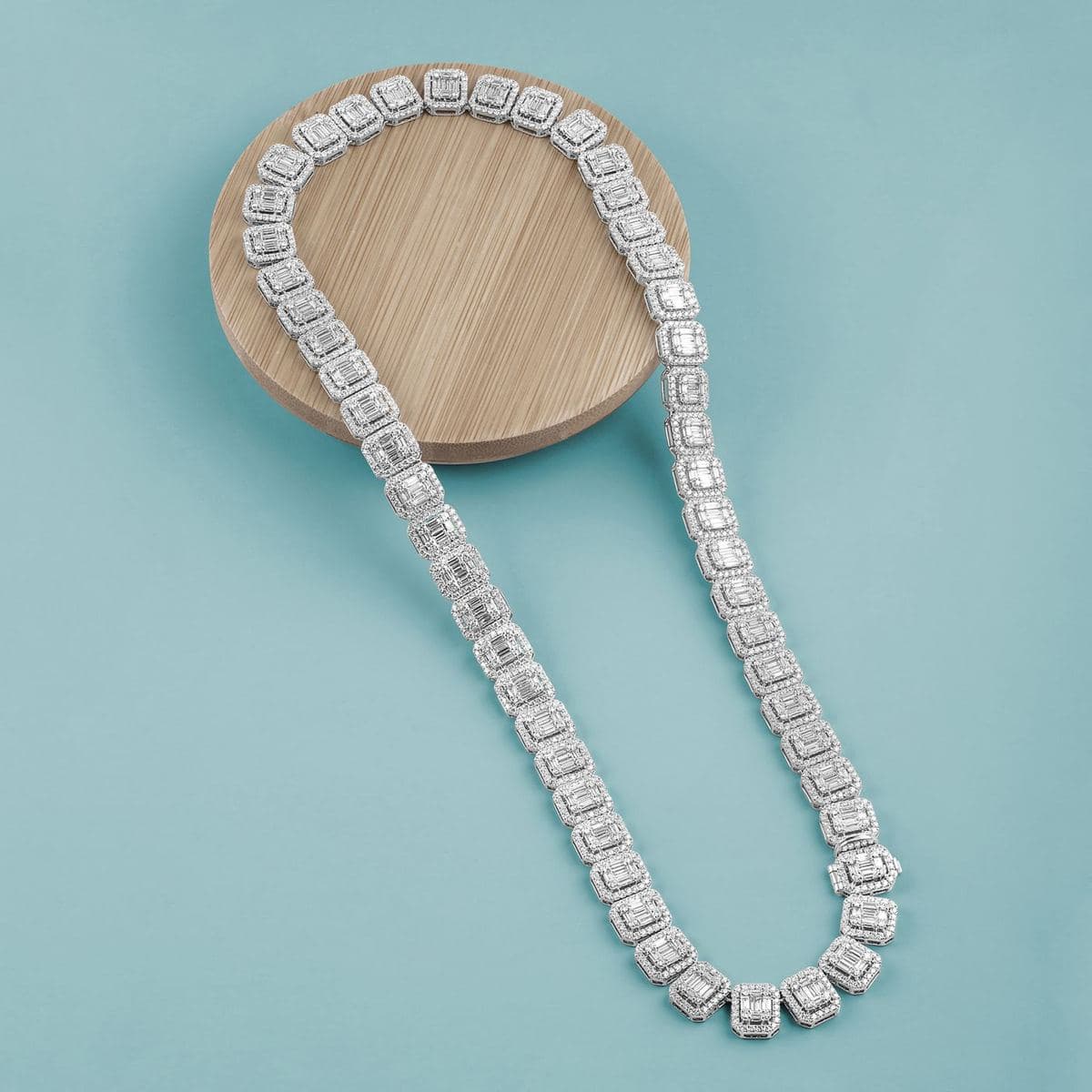 Flat-lay of a white-metal tennis chain made of uniform rectangular emerald-shaped baguette-cluster links each framed by a halo of small round stones, looped over a wooden disc