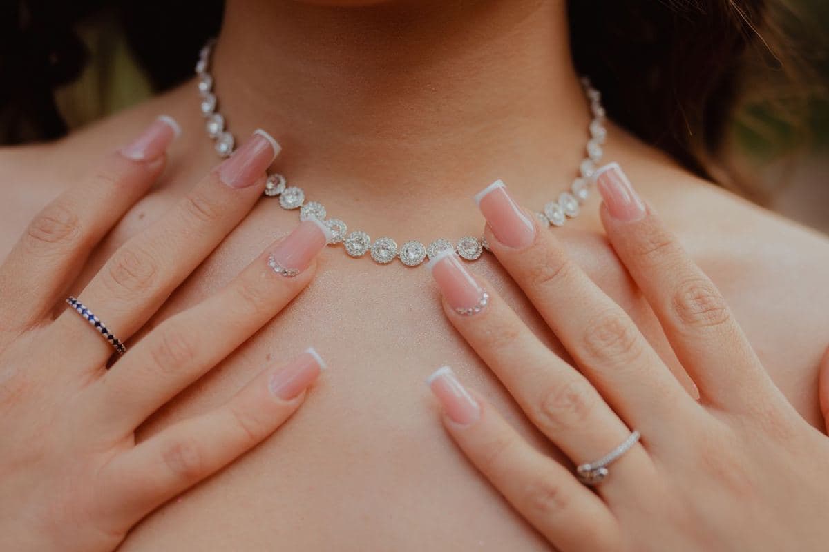 Close-up of a neck with a line of round white stones each in a small-stone halo, white metal, framed by two hands with French manicure and rings