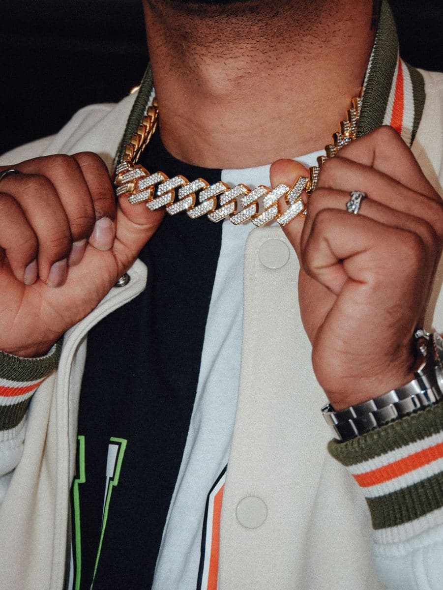 Man, face cropped above the chin, holding a heavy yellow-gold cuban link chain whose links are paved with small white stones, wearing a steel watch