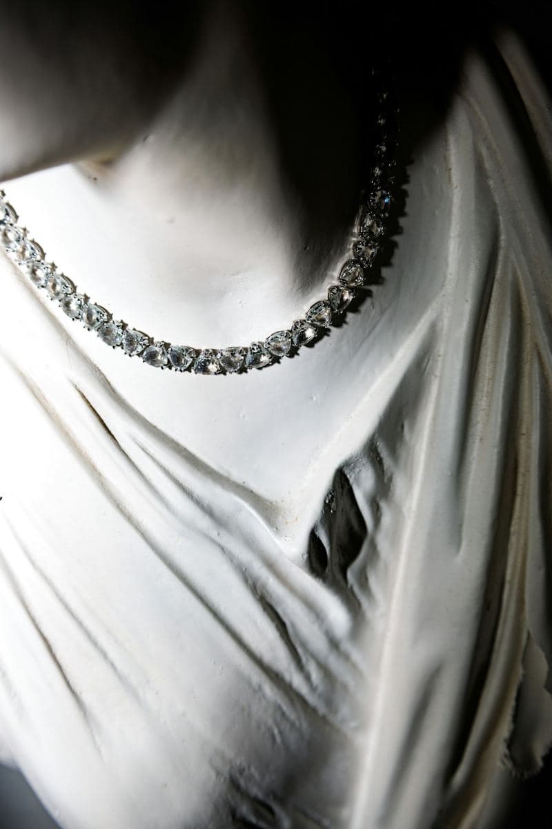A single line of prong-set pear-shaped clear stones in white metal worn around the neck of a white classical statue