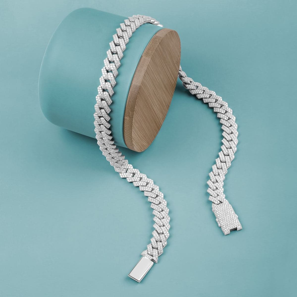 Flat-lay of a white-metal prong-style cuban link chain fully paved with small round stones, draped over a teal cylinder, pavé box clasp visible